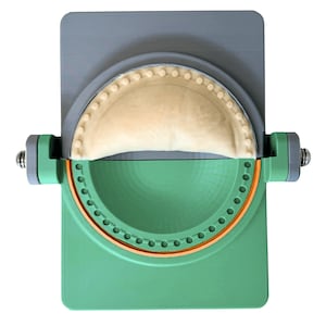 May include: A green and gray empanada maker with a half-moon shape. The device has a gray top, a green base, and a circular orange accent. A filled empanada is visible inside the mold, ready for sealing. The tool is designed for easy empanada preparation.