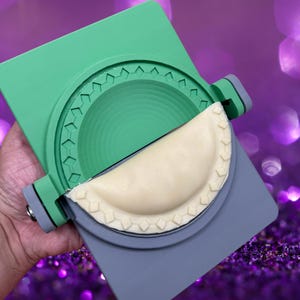 Pode incluir: Um molde de plástico verde e cinza para fazer empanadas. O molde tem um design texturizado e uma tampa com dobradiças. Uma empanada a meio fazer está no molde.