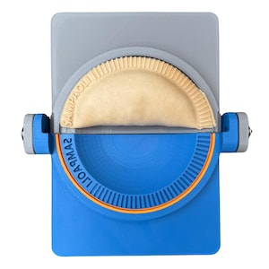 May include: A blue and gray empanada maker with a half-moon shaped dough inside. The tool has a blue base with the text "UOAGMA2" and an orange accent. The top is gray with the text "SAMPAOLI".