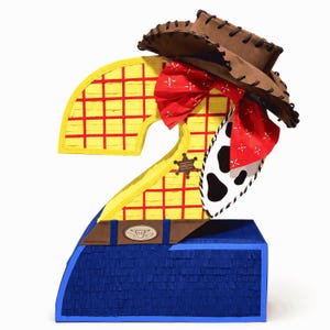 Puede incluir: Una piñata con forma de número dos que presenta un patrón de cuadrícula amarillo, un sombrero de vaquero marrón, un pañuelo rojo con detalles blancos, una sección con estampado de vaca, un cinturón marrón y una base azul oscuro.