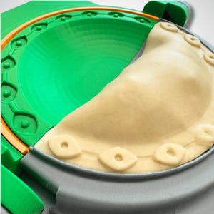May include: A green and gray plastic dumpling press with a circular design, featuring a half-moon shaped mold. A piece of beige raw dough is pressed into the mold, showing a decorative embossed pattern along the curved edge of the pastry.