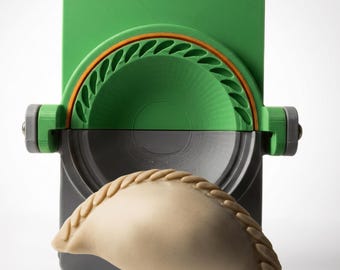Puede incluir: Una prensa para empanadillas de plástico verde y gris con diseño articulado y borde semicircular para sellar. Una masa de empanadilla beige sin hornear con un borde trenzado decorativo se sitúa delante, mostrando el patrón del molde.
