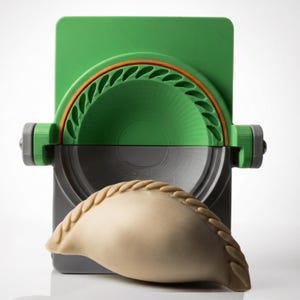 Peut inclure: Une presse à raviolis en plastique vert et gris dotée d'un mécanisme à charnière avec un bord de sertissage semi-circulaire. Une pâte à ravioli beige non cuite avec une couture tressée décorative est placée devant l'outil de cuisine.