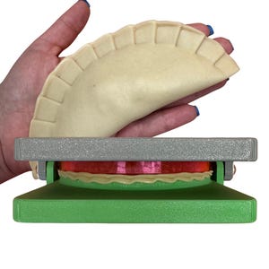 May include: A green and gray plastic empanada press with a red and white silicone ring. The press is open and a hand is holding a raw empanada above the press.