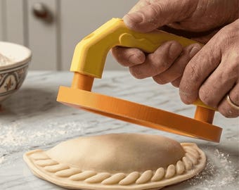 Puede incluir: Una prensa de empanadas naranja y amarilla se utiliza para dar forma a una masa rellena. La prensa tiene un mango y una base circular, y se presiona sobre la masa. La masa es de color marrón claro con un borde decorativo.
