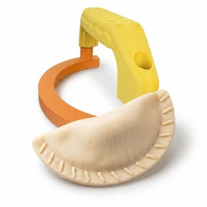 May include: A yellow and orange ravioli maker with a semi-circular orange base and a yellow handle. A beige, uncooked ravioli is in the foreground. The ravioli maker is designed to shape and seal pasta.