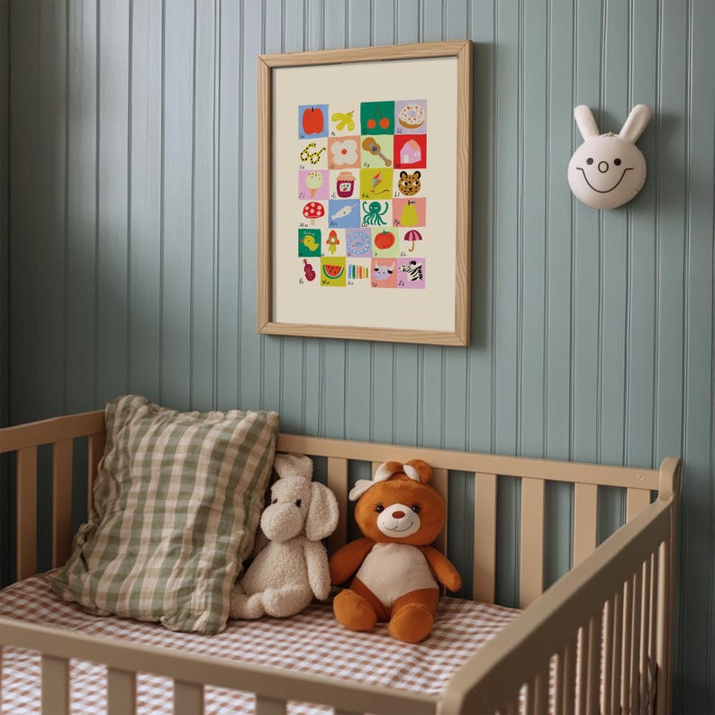 May include: A framed alphabet art print with colorful illustrations hangs on a blue wall above a crib. The print features a variety of images, including an apple, a donut, and a tiger. A white bunny head wall decoration is also visible.