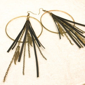 May include: A pair of gold hoop earrings with black leather fringe and gold chain accents. The earrings have a bohemian style.