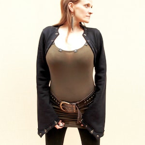 Puede incluir: Bolero negro con remaches metálicos y mangas largas. Se lleva sobre un top verde oscuro y un cinturón marrón con tachuelas y una hebilla grande. El conjunto se completa con leggings negros.