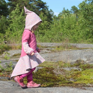 May include: A young child wearing a pink hooded jacket with a colourful pattern. The jacket has a long, flowing skirt and the child is walking on a rocky surface.