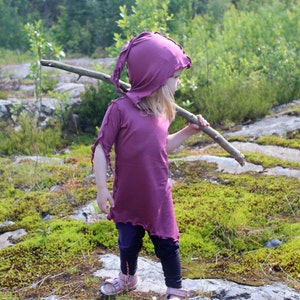 May include: A young person wearing a purple hooded tunic and black leggings holds a stick in a forest setting.