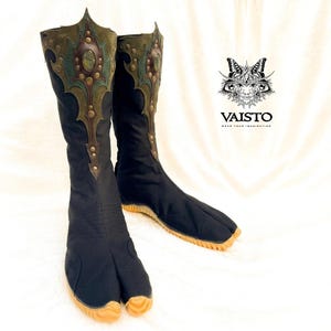 May include: A pair of black split-toe boots with a tan rubber sole. The upper shaft features an olive green and dark brown leather overlay with brass studs and a central oval stone. The logo VAISTO with a butterfly graphic is visible.
