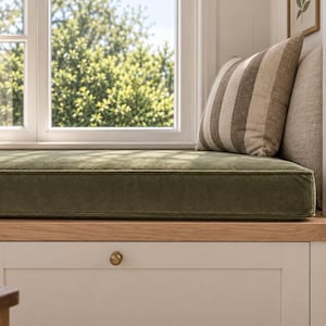 Può includere: Un sedile da finestra con un cuscino spesso in velluto verde oliva posizionato su una panca in legno di rovere chiaro con mobiletti bianchi sottostanti. Un cuscino a righe beige e marrone poggia sul sedile, contro un telaio bianco.