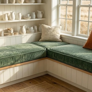 May include: A comfortable window seat with green velvet cushions and a beige throw. The built-in bench has a wooden base and is complemented by neutral and rust-coloured pillows. White shelves display ceramic dishware.