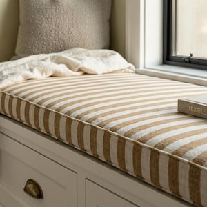May include: A white window bench with drawers featuring brass cup pulls, topped with a tan and white striped cushion. A beige textured pillow, a white fabric throw, and a book titled TEXTILE DESIGN rest on the bench near a window.