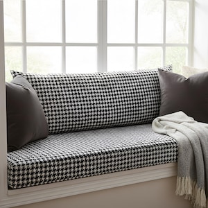 May include: A window seat featuring a black and white houndstooth cushion and backrest. Dark grey and cream-coloured cushions are arranged against the backrest. A grey throw blanket with fringe and a white mug on a small wooden table.