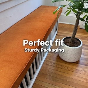 Op de afbeelding: Een oranje bankkussen met de tekst "Perfect fit" en "Sturdy Packaging" is geplaatst op een witte lattenbodem. Een potplant met een gevlochten stam staat in de buurt op een hardhouten vloer.