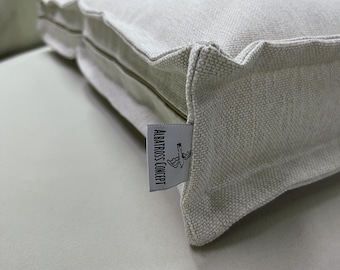 May include: Close-up of a rectangular, light gray cushion with a textured fabric. The cushion has a piped edge and a tag that reads "ALBATROSS CONCEPT" with a small illustration. The cushion is resting on a light gray surface.
