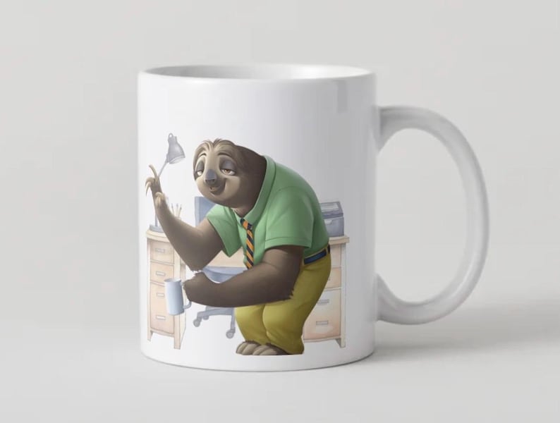 Op de afbeelding: Witte keramische mok met een cartoon luiaard karakter in een groen shirt en gele broek, met een kleine mok. De luiaard bevindt zich in een kantooromgeving met een bureau en een lamp. De mok heeft een wit handvat en een wit interieur.