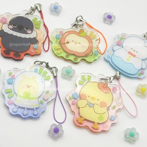 May include: A collection of clear acrylic charms with cartoon character designs, including a bird, apple, alien, and chick. Each charm has a colorful border and a lanyard. The charms are decorated with floral accents and the text "@sparkofapple".