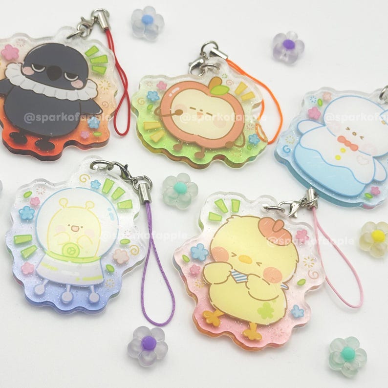 May include: A collection of clear acrylic charms with cartoon character designs, including a bird, apple, alien, and chick. Each charm has a colorful border and a lanyard. The charms are decorated with floral accents and the text "@sparkofapple".