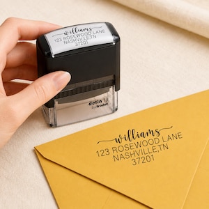 Op de afbeelding: Een zwarte zelfinktende stempel met een doorzichtige basis, voorzien van de tekst Williams 123 Rosewood Lane Nashville, TN 37201. De stempel ligt naast een gele envelop met hetzelfde adres in zwarte inkt, afgedrukt door het stempelapparaat.