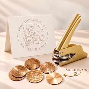 May include: A gold-colored metal embosser tool with a lever mechanism, accompanied by several circular brass-colored metal discs. A white card features an embossed design of a skeleton reading a book with the text FROM THE LIBRARY OF NATALEE PEER.