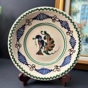 May include: A round ceramic plate with a cream-coloured base featuring a hand-painted blue and orange bird in the centre. The rim is decorated with a green zigzag border and alternating blue wavy lines and brown scroll motifs around the edge.