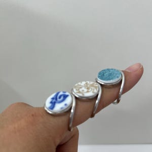 May include: Three silver-toned rings featuring circular ceramic inlays. The first ring has a white inlay with blue patterns, the second has a white inlay with gold swirls, and the third has a textured, solid teal-colored inlay.
