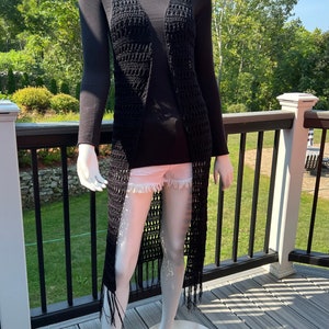 Può includere: Un lungo gilet di uncinetto nero con dettagli a frange. Il gilet è aperto sul davanti e ha una vestibilità ampia.