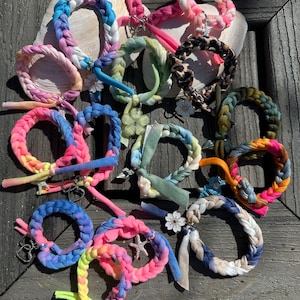 May include: A collection of colorful braided bracelets with various charms, including hearts, stars, and flowers. The bracelets are made from tie-dye fabric and are arranged on a wooden surface.