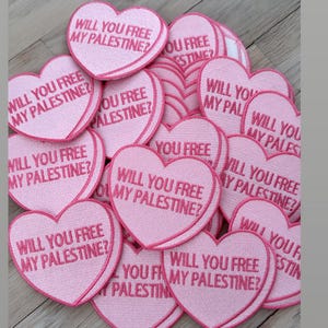 May include: A pile of pink heart-shaped patches with the text "Will you free my Palestine?" on them. The patches are made of felt and have a pink embroidered border.