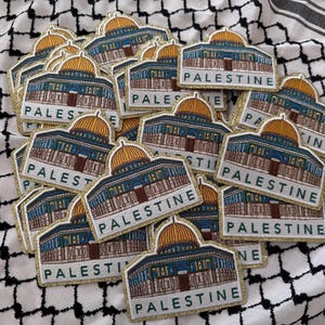 Puede incluir: Un conjunto de parches bordados con la Cúpula de la Roca y la palabra "PALESTINA" en letras blancas. Los parches tienen un borde dorado y están colocados sobre una tela a cuadros en blanco y negro.