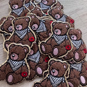 May include: Brown teddy bear patches with gold glitter trim, wearing a black and white bandana and a red flower. The patches are embroidered with pink accents on the paws and ears. The bears are piled together.