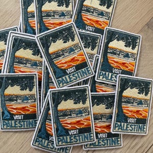 May include: A collection of embroidered patches with a rectangular design. Each patch features a tree, a landscape, and the words "VISIT PALESTINE" in white. The patches have a white border and are arranged on a wooden surface.