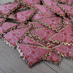 May include: A collection of red and white patterned fabric triangles with gold scalloped edges. The fabric has a woven design with red lines and a gold trim. The triangles are arranged on a light wood surface.