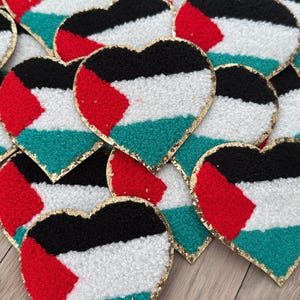 May include: A pile of heart-shaped patches with the Palestinian flag colors: black, white, red, and green. The patches have a gold glitter border.