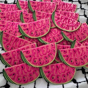Puede incluir: Parches bordados de sandía en tonos rosa y verde, con el texto "FREE CESS". Los parches están dispuestos sobre una tela a cuadros en blanco y negro.