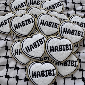 May include: A collection of heart-shaped patches with the word "HABIBI" embroidered in black on a white background. Each patch has a gold border and is arranged on a black and white checkered fabric.
