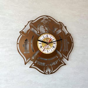 Puede incluir: Un reloj de pared de metal marrón con forma de cruz de Malta de bombero. La esfera del reloj tiene números romanos y manecillas negras. Las palabras "FIRE DEPT" son visibles, junto con un símbolo de llama.