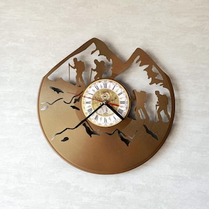 May include: A circular wall clock featuring a bronze-colored metal cutout of mountain peaks and hikers. The center displays a round clock face with white background, black Roman numerals, and black hour and minute hands with a red second hand.