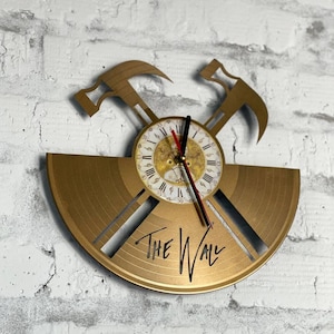 May include: A gold-colored wall clock made from a vinyl record. The clock features a clock face with Roman numerals and two hammer accents. The words "THE WALL" are cut into the record. The clock is mounted on a white brick wall.