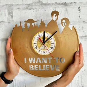 May include: A gold-colored vinyl record clock with the words "I WANT TO BELIEVE" in silver lettering. The clock features a forest silhouette, a UFO, and two figures. The clock hands are black with a red second hand.