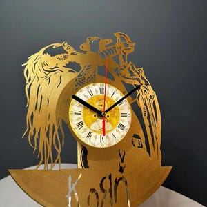 May include: A gold-colored decorative clock with a circular face featuring Roman numerals and black hands. The clock is cut from a vinyl record and has a decorative design with the word "Kosin" cut out at the bottom.