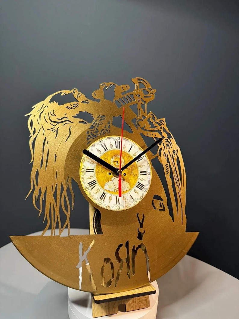 May include: A gold-colored decorative clock with a circular face featuring Roman numerals and black hands. The clock is cut from a vinyl record and has a decorative design with the word "Kosin" cut out at the bottom.