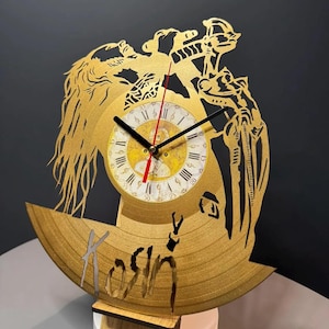 May include: A gold-colored vinyl record clock with a cut-out design featuring a figure and the word "Kosin". The clock face has Roman numerals and black hands with a red second hand. The clock is mounted on a wooden stand.