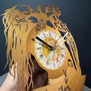 May include: Gold-colored wall clock crafted from a repurposed vinyl record. The clock features a detailed, laser-cut design with an ornate face displaying Roman numerals and decorative hands. The clock's diameter is approximately 12 inches.