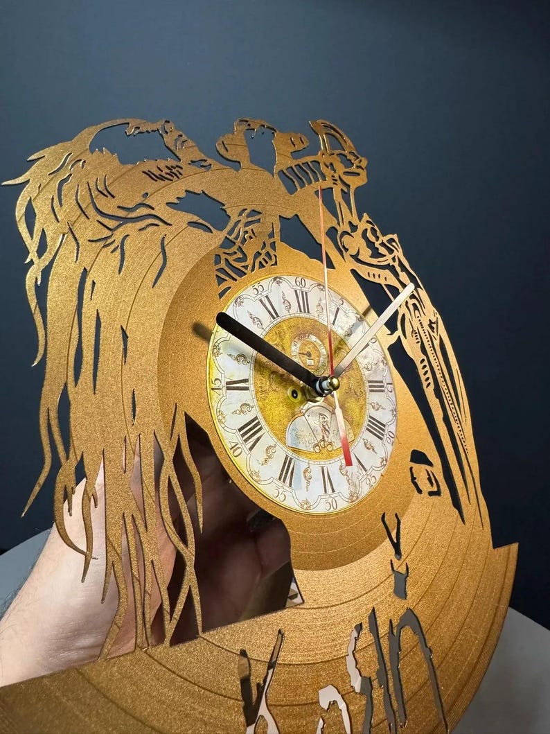May include: Gold-colored wall clock crafted from a repurposed vinyl record. The clock features a detailed, laser-cut design with an ornate face displaying Roman numerals and decorative hands. The clock's diameter is approximately 12 inches.