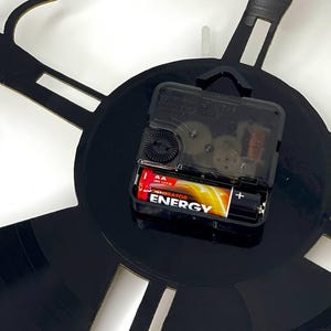 May include: A black wall clock with a unique design, possibly a musical theme. The clock mechanism is visible, powered by a AA battery. The clock hands are not visible. The clock is mounted on a white wall.
