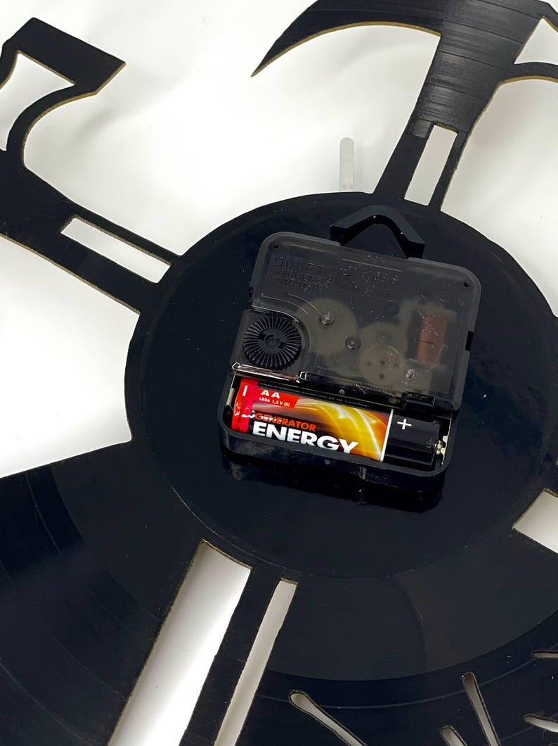 May include: A black wall clock with a unique design, possibly a musical theme. The clock mechanism is visible, powered by a AA battery. The clock hands are not visible. The clock is mounted on a white wall.